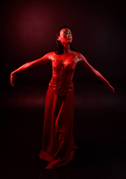 Πλήρες πορτρέτο της όμορφης νεαρής Ασιάτισσας που φοράει κόκκινο κορσέ, καλυμμένο με ματωμένη μπογιά που ποζάρει σε γοτθικό ύφος τρόμου με δημιουργικές χειρονομίες απομονωμένες σε σκούρο μαύρο φόντο στούντιο. - Φωτογραφία, εικόνα
