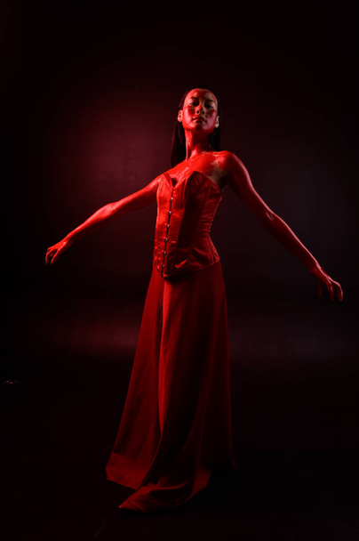 Full length portrait of beautiful young asian woman wearing red corset, covered with bloody paint posing in gothic horror style with creative hand gestures isolated against dark black studio background. - Fotografie, Obrázek