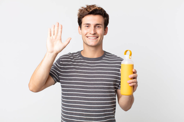 young handsome man smiling happily, waving hand, welcoming and greeting you and holding a coffee thermos - Foto, afbeelding