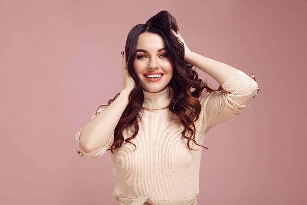 Attractive caucasian toothy smiling woman touching her clean brunette hair and expresses pleasure standing isolated over pink background - Photo, Image