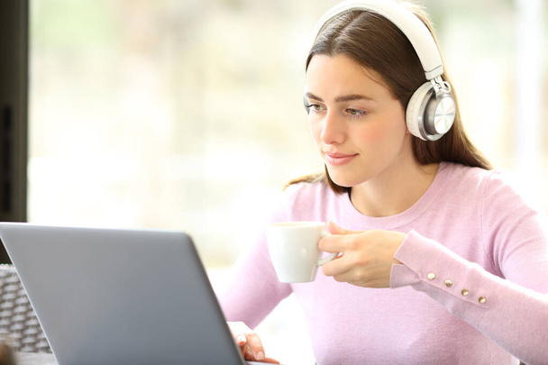 Relaxed woman using laptop and wireless headphones drinking coffee in a bar - Фото, изображение