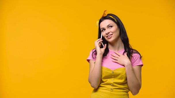 Мечтательная женщина в повязке говорит на смартфоне изолированы на желтый - Фото, изображение