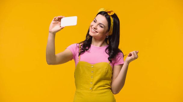 Smiling woman in headband taking selfie on smartphone isolated on yellow - Фото, зображення