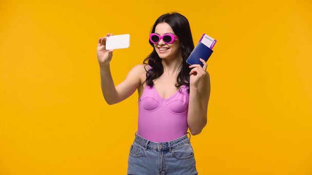 Młoda kobieta w okularach przeciwsłonecznych robi selfie i trzyma paszport odizolowany na żółto - Zdjęcie, obraz