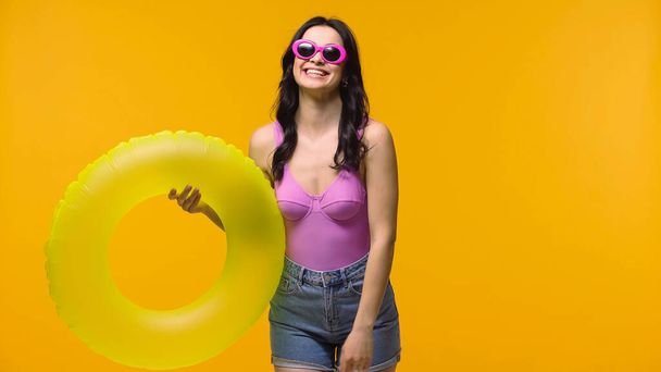 Χαρούμενη γυναίκα με γυαλιά ηλίου που κρατά το δαχτυλίδι κολύμβησης απομονωμένο στο κίτρινο  - Φωτογραφία, εικόνα