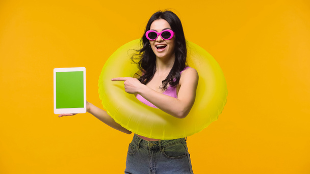 Femme excitée en lunettes de soleil et anneau de natation pointant vers comprimé numérique isolé sur jaune  - Photo, image