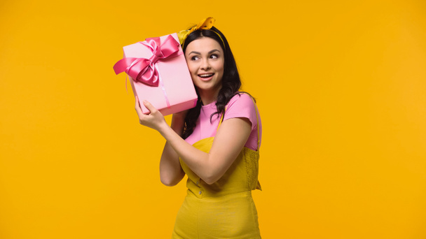 Positive woman holding present near ear isolated on yellow  - Foto, afbeelding