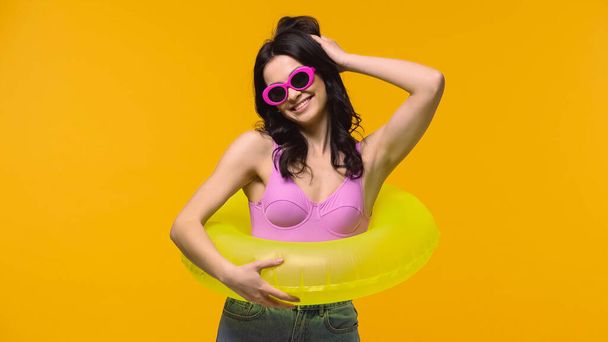 Smiling woman in sunglasses and swim ring adjusting hair isolated on yellow  - Photo, Image
