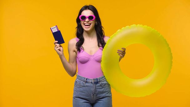 Excited woman in sunglasses holding passport with air ticket and swim ring isolated on yellow  - 写真・画像
