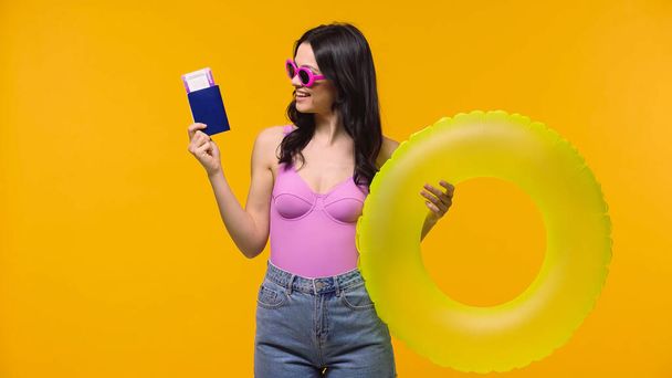 Вид збоку красива жінка в сонцезахисних окулярах дивиться на паспорт, тримаючи плавальний кільце ізольовано на жовтому
  - Фото, зображення