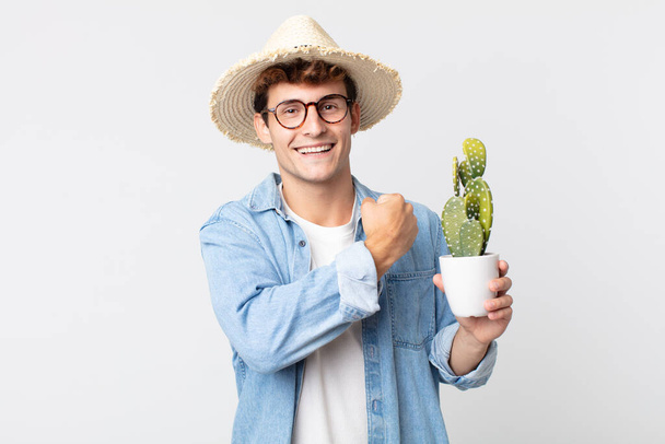 young handsome man feeling happy and facing a challenge or celebrating. farmer holding a decorative cactus - Foto, afbeelding