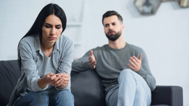 worried woman sitting and looking at hands near blurred and displeased boyfriend quarrelling in living room - Photo, Image