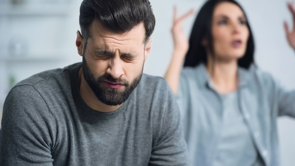 smutný a vousatý muž se zavřenýma očima poslouchající rozmazané přítelkyně hádající se doma - Fotografie, Obrázek