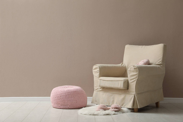 Knitted pouf, fuzzy rug with slippers and armchair near beige wall indoors. Space for text - Фото, изображение