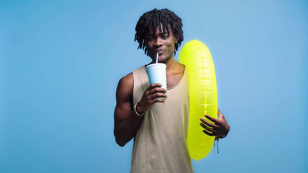 African american man with paper cup drinking soft drink and holding inflatable ring isolated on blue - Photo, Image