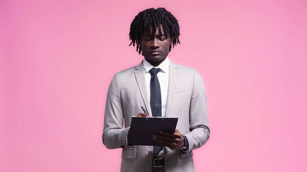 african american man in suit writing on clipboard isolated on pink - Photo, Image