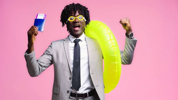 Homme d'affaires afro-américain avec billet d'avion et anneau gonflable se réjouissant isolé sur rose - Photo, image