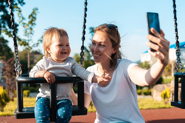 若い白人の幸せな女性の肖像画はスイングに座って幼児と自撮りを取ります.背景に遊び場。夏. - 写真・画像