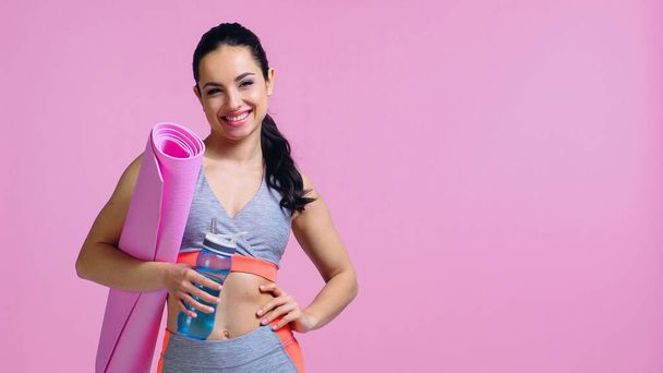 vrolijke sportvrouw houden sportfles en fitness mat terwijl staan met de hand op heup geïsoleerd op roze - Foto, afbeelding