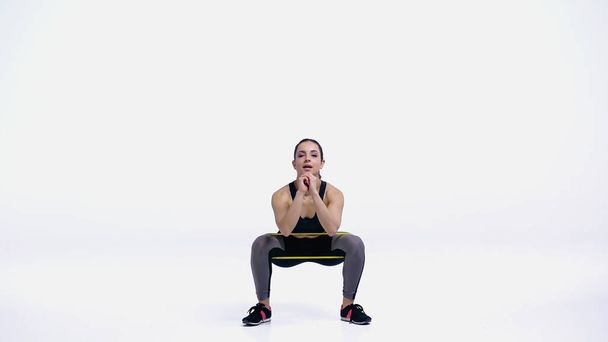 brunette jonge sportvrouw doet squat met geel elastiek op wit - Foto, afbeelding