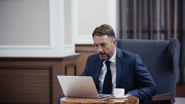Businessman in suit using laptop near cup of coffee in lobby of restaurant - Фото, зображення