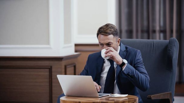 Businessman in suit drinking coffee and using laptop in lobby of restaurant - Foto, afbeelding