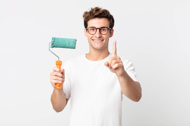 young handsome man smiling proudly and confidently making number one and holding a roller paint - Fotoğraf, Görsel