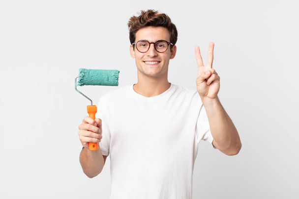 young handsome man smiling and looking friendly, showing number two and holding a roller paint - Fotografie, Obrázek