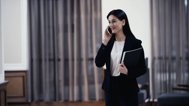 cheerful businesswoman with paper folder talking on smartphone in lobby of restaurant - Valokuva, kuva