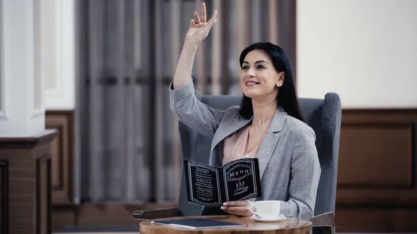 cheerful businesswoman holding menu and gesturing in lobby of restaurant - Foto, Imagem