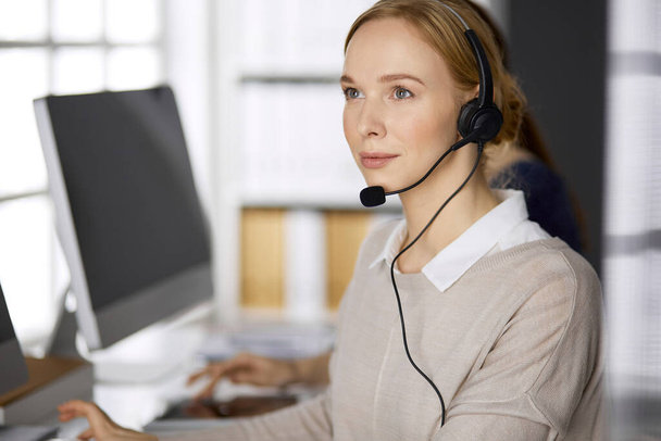 Friendly businesswoman talking by headset while sitting in office. Call center and diverse people group in business - Фото, изображение