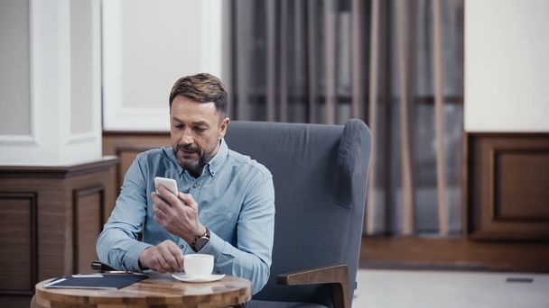 Businessman using smartphone and reaching cup of coffee in lobby of restaurant - Foto, Imagem