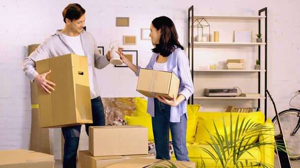 smiling man giving statuette to happy woman while standing with boxes in new home  - Photo, Image