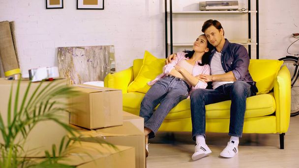 Smiling couple talking and resting on couch near boxes in new home - Photo, Image