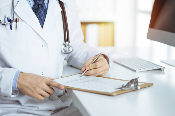 Friendly red-bearded doctor sitting and writing with clipboard in clinic, close-up - Photo, Image