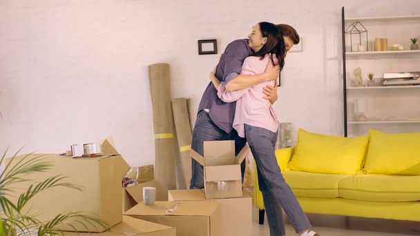 happy couple hugging near carton boxes in new home - Фото, изображение