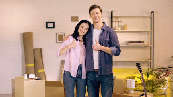 Couple souriant montrant pouces près des boîtes dans la nouvelle maison - Photo, image