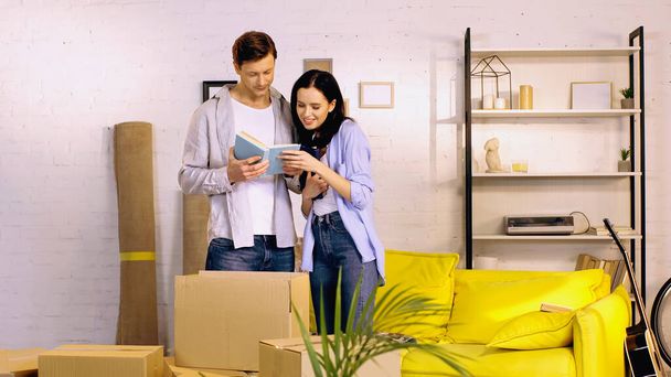 cheerful young couple reading book near boxes in living room - Photo, Image