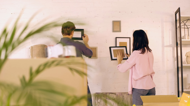 vista trasera de la pareja colgando marcos de fotos en la pared cerca de cajas en un nuevo hogar - Foto, Imagen