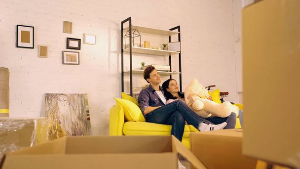 cheerful couple chilling on sofa with teddy bear near boxes in new home  - Photo, Image