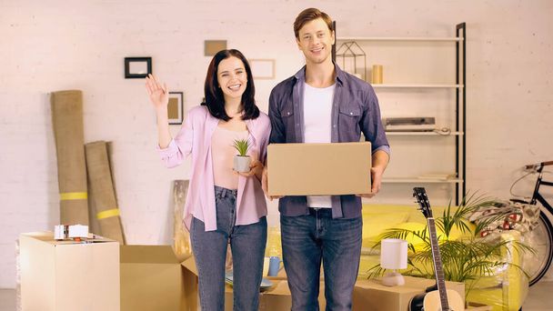 happy woman waving hand and holding plant near man with box in new home - 写真・画像