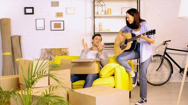 young woman playing guitar near boyfriend applauding in new home - Foto, Imagen