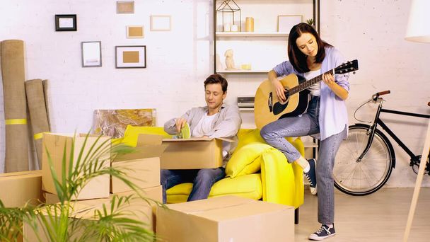 young woman playing guitar near boyfriend unpacking box in new home - 写真・画像