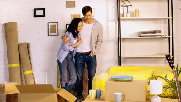 happy man standing with hand in pocket and hugging cheerful girlfriend in new home - 写真・画像
