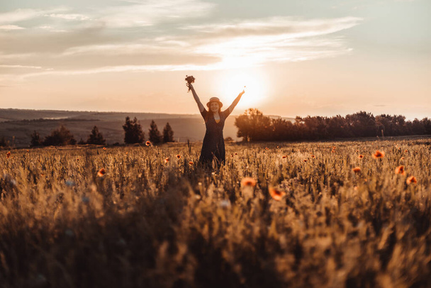 Caucasian Happy beautiful young girl with a poppy bouquet in her hands in a summer field at sunset. Tourism, traveling and healthy lifestyle concept. - Photo, Image