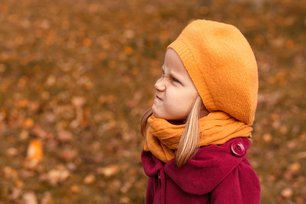 Klein meisje in een gele baret is boos. Portret van een kind in herfstkleding op de achtergrond van een glade met gevallen bladeren - Foto, afbeelding