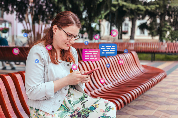 Съемка улыбающейся молодой женщины с мобильного телефона, чтение сообщений на мобильном телефоне в городском парке - Фото, изображение