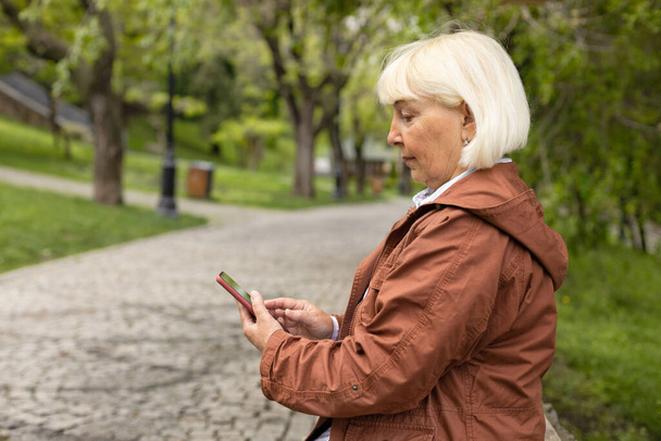 Aktif yetişkin bir kadın parkta bir bankta otururken akıllı telefon kullanıyor. - Fotoğraf, Görsel