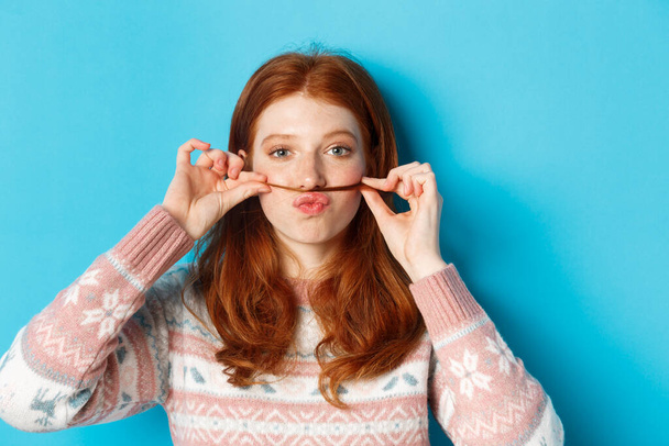 Close-up of silly and funny redhead girl making moustache with hair strand and puckered lips, grimacing against blue background - Photo, Image