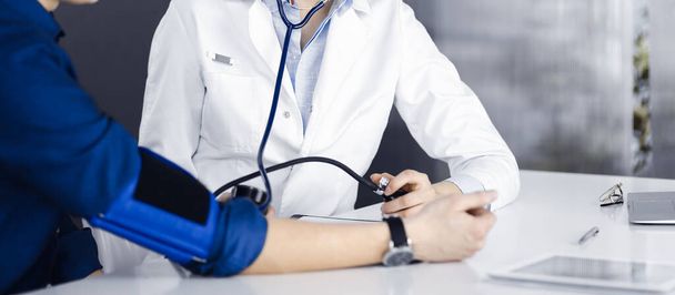 Médica desconhecida está verificando a pressão arterial de seus pacientes enquanto estão sentados juntos na clínica. Conceito de medicina durante a pandemia de Coronavirus. Covid 2019 - Foto, Imagem